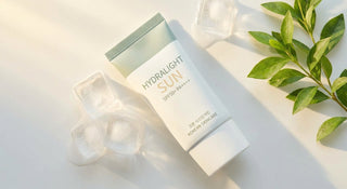 A minimalist flat-lay of a Korean sunscreen tube surrounded by ice cubes and green leaves in warm summer sunlight on a clean white surface.