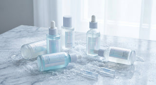 Minimalist Korean skincare ampoules and serums arranged on a frosted marble surface in cool pearl and icy blue tones, glowing under soft natural light.